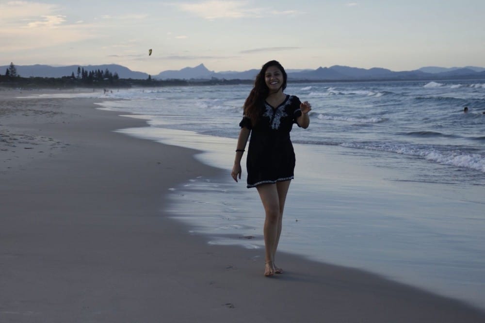 chica morena con un vestido negro andando por la orilla de la playa descalza y sonriente